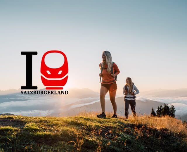 Zwei Wanderer auf einem grasbewachsenen Hügel bei Sonnenaufgang mit dem I love SalzburgerLand-Logo auf der linken Seite.