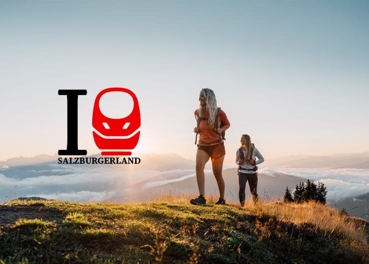 DB-iBahn-teaser Zwei Wanderer auf einem grasbewachsenen Hügel bei Sonnenaufgang mit dem I love SalzburgerLand-Logo auf der linken Seite.