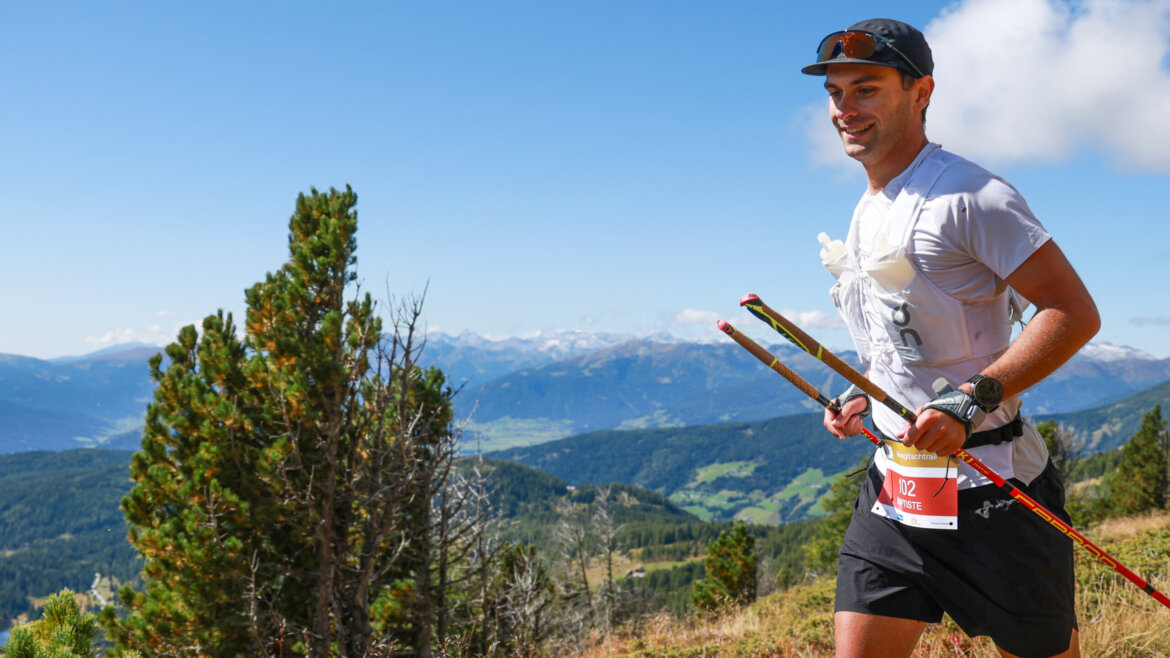 Mann beim Trailrunning mit Trekkingstöcken auf einem Bergpfad, mit Startnummer, malerische Hügel im Hintergrund. (vergrößerte Ansicht)