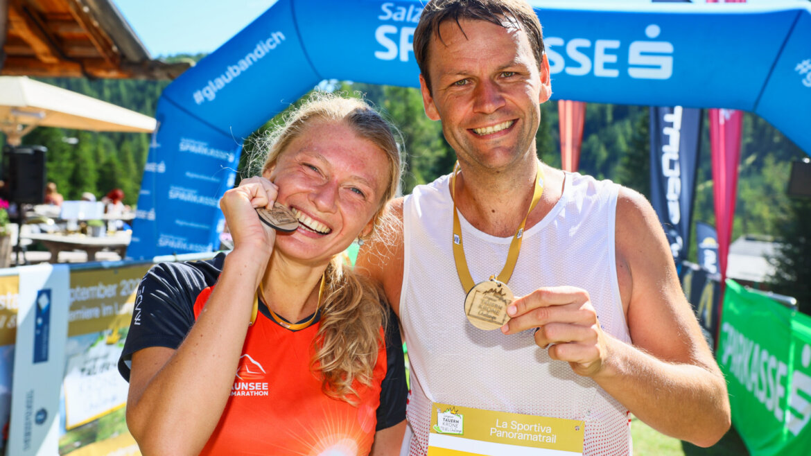 Zwei lächelnde Läufer zeigen ihre Trailrunning-Medaillen an einer Ziellinie mit bunten Veranstaltungsbannern im Hintergrund. (vergrößerte Ansicht)