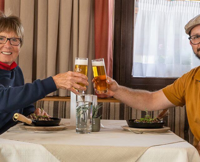 Zwei Menschen sitzen an einem Tisch in einem Restaurant, lächeln und stoßen mit Biergläsern an.