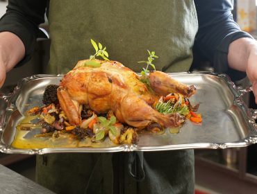 SalzburgerLand schmecken und entdecken mit Rudi Pichler im Tennengau Person mit einem gebratenen Huhn, garniert mit Kräutern und Gemüse auf einem Silbertablett in einer Küche.