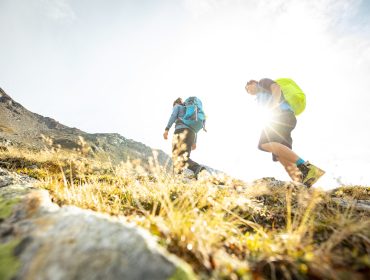 Zwei Personen mit Rucksäcken genießen ein Bike & Hike-Abenteuer an einem grasbewachsenen Berghang im hellen Sonnenlicht.