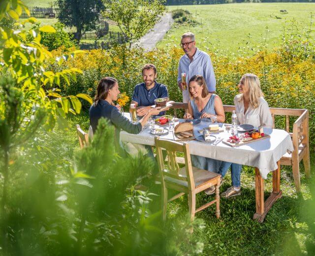 Fünf Personen genießen eine Mahlzeit an einem weißen Tisch in einem sonnigen, grünen Garten, in dem Nachhaltigkeit und Natur groß geschrieben werden.