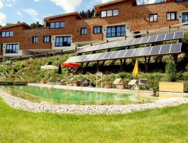 Bachalm Sonnenkollektoren an einem Hang über einem natürlichen Swimmingpool und Liegestühlen, mit Holzgebäuden im Hintergrund.