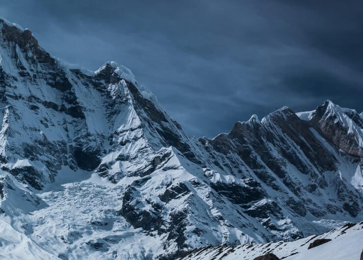 Schneebedeckte Berggipfel unter einem dunklen, wolkenverhangenen Himmel mit felsigen Hängen und einer dramatischen Landschaft.