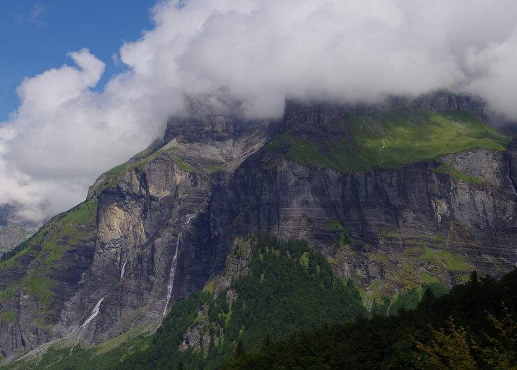 Wolken bedecken teilweise steile, felsige grüne Berge mit einem Waldgebiet im Vordergrund.