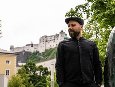 Ein bärtiger Mann in schwarzer Kleidung steht im Freien in der Nähe einer Statue, im Hintergrund ist ein Schloss auf einem Hügel zu sehen.