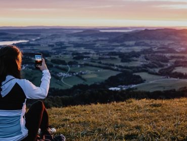 Eine Person, die bei Sonnenuntergang auf einem Hügel sitzt und mit ihrem Smartphone ein Foto von der malerischen Landschaft macht.