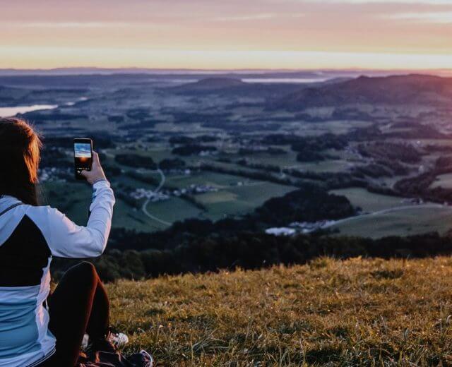 Eine Person, die bei Sonnenuntergang auf einem Hügel sitzt und mit ihrem Smartphone ein Foto von der malerischen Landschaft macht.