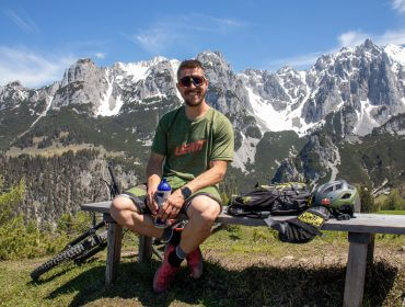 Mann sitzt auf einer Bank mit Fahrradausrüstung, lächelnd, Berge mit Schnee im Hintergrund, sonniger Tag.