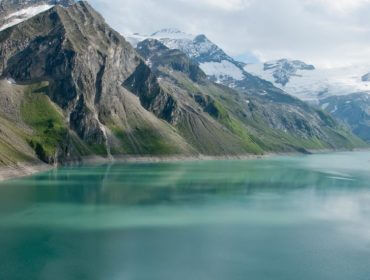 Ein türkisfarbener Stausee, umgeben von steilen, felsigen Bergen und schneebedeckten Gipfeln unter einem teilweise bewölkten Himmel.