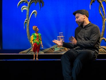 Scheibsta im Salzburger Marionettentheater Ein Mann in Schwarz spricht mit einer farbenfrohen Puppe, mit Palmenrequisiten und einem blauen Hintergrund auf der Bühne.