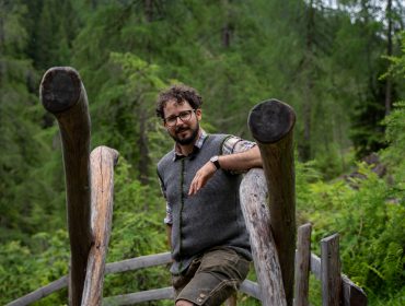 Ein Mann mit Brille lehnt an einem rustikalen Holzzaun in einem üppigen, grünen Wald.