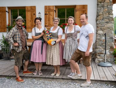 Rudi‘s Roadtrip durch das SalzburgerLand – Almroas Edition HaibenAlm St. Johann Alpendorf Fünf lächelnde Erwachsene in traditioneller österreichischer Tracht posieren vor einem Haus mit einer bunten Kuhglocke.