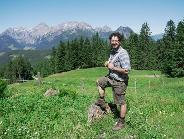 Ein Mann in Wanderkleidung lächelt, während er auf einem Baumstumpf in einer grünen Wiese mit Bergen und Bäumen im Hintergrund steht.
