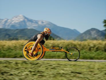Eine Person fährt mit einem Handbike auf einer Straße mit Bergen und Feldern im Hintergrund.