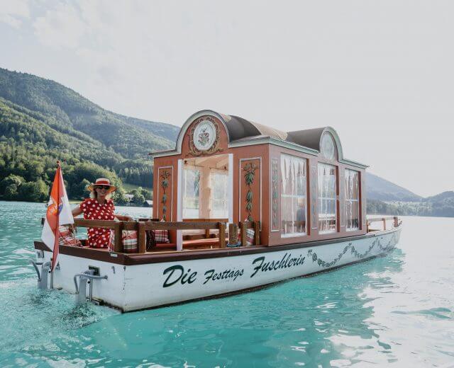 Eine Frau in einem roten Kleid steht auf einem geschmückten Hausboot mit Schweizer Flaggen auf einem türkisfarbenen See.