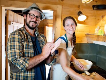 Ein lächelnder Mann und eine lächelnde Frau bereiten in einer gemütlichen Holzküche gemeinsam Essen zu.