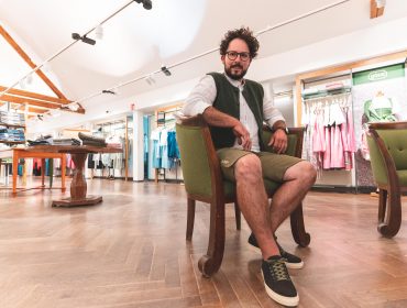 Rudi‘s Roadtrip durch das SalzburgerLand – Bauernherbst Edition, Gössl Trachten Ein Mann mit Brille sitzt auf einem grünen Stuhl in einem Bekleidungsgeschäft mit Holzboden und Kleiderständern.