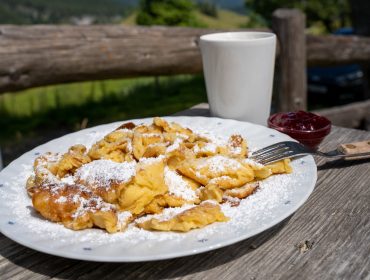 Teller mit Puderzucker bestreuten Pfannkuchenstücken mit einer Gabel, Marmelade und einem weißen Becher auf einem rustikalen Holztisch.