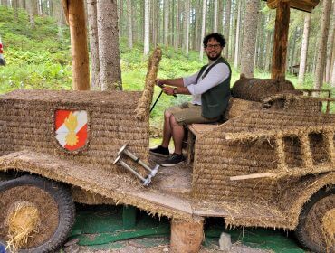 Ein Mann lächelt, während er in einem rustikalen Auto aus Heuballen in einer Waldlandschaft sitzt.