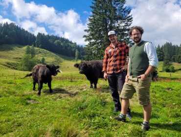 Zwei Männer stehen lächelnd in einem grünen Faistenauer Feld mit Kühen, Hügeln, Bäumen und blauem Himmel im Hintergrund.