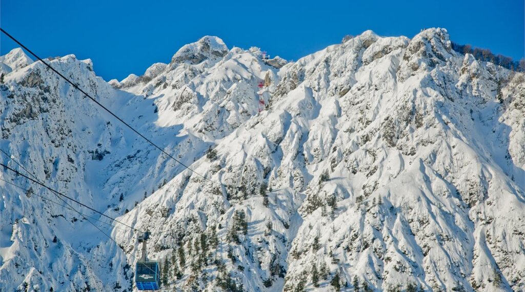 Eine blaue Seilbahn erklimmt schneebedeckte, zerklüftete Berggipfel unter einem strahlend blauen Himmel.