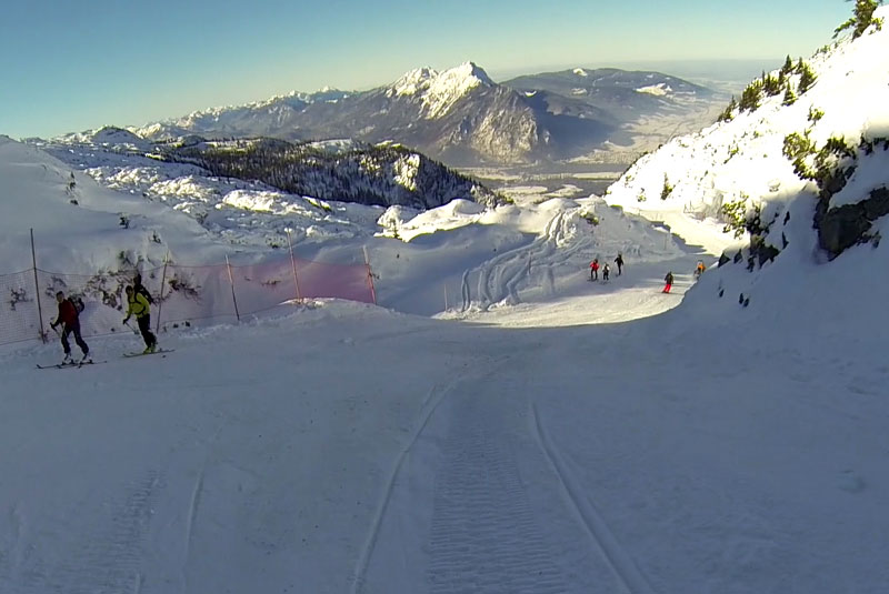 Skifahrer auf einem verschneiten Berghang mit fernen Bergen und einem klaren Himmel im Hintergrund.