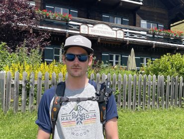 Ein Mann mit Sonnenbrille und Mütze steht vor einem rustikalen Holzhaus mit Blumen und einem Lattenzaun.