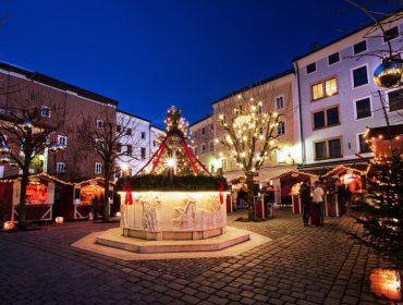 Ein festlicher Stadtplatz bei Nacht mit Weihnachtsbeleuchtung, geschmückten Bäumen und Marktständen rund um einen Brunnen.