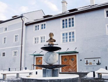 Schneebedeckter Hof mit rundem Brunnen und Büstenstatue vor einem hellgrauen Gebäude mit vielen Fenstern.