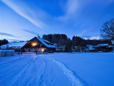 Ein verschneites Bauernhaus erstrahlt in der Abenddämmerung in warmem Licht, umgeben von Bäumen und Bergen.