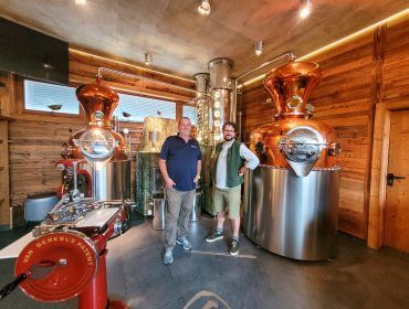 Rudi‘s Roadtrip durch das SalzburgerLand – Bauernherbst Edition, Pinzgau Zwei Männer stehen lächelnd in einer Destillerie mit kupfernen Brennblasen und holzgetäfelten Wänden im Hintergrund.