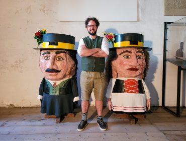 Ein Mann steht zwischen zwei großen Puppen mit bemalten Gesichtern, Hüten und bunten Kleidern in einem Raum auf einem Holzboden.