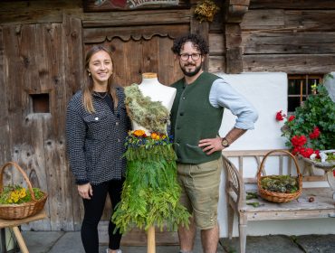 Zwei Personen stehen neben einer Schaufensterpuppe, die ein von der Natur inspiriertes Kleid trägt, vor einem rustikalen Holzgebäude.