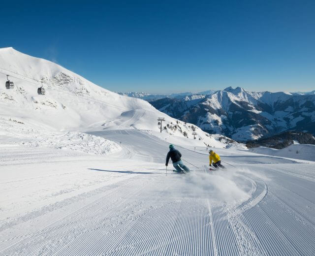 Zwei Skifahrer gleiten eine präparierte Schneepiste hinunter, die Berge und Skilifte hinter sich lassend, die Nachhaltigkeit.