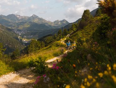 Zwei Wanderer genießen das Wandern im Almherbst auf einem Weg zwischen grünen Hügeln, Bergen und einem entfernten Dorf.