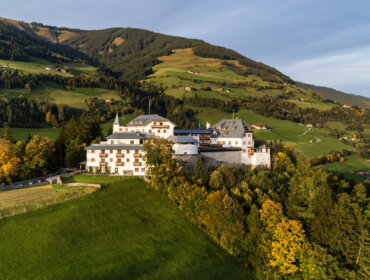 Luftaufnahme von Schloss Mittersill, einem großen Schloss am Hang inmitten von Bäumen und sanften grünen Hügeln unter blauem Himmel.