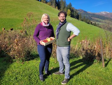 Zwei Personen stehen auf einem grasbewachsenen Hügel; eine hält eine Kiste mit Äpfeln. Im Hintergrund sind Bäume und Berge zu sehen.