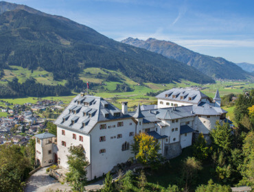 Schloss Mittersill liegt auf einem grünen Hügel, mit Blick auf ein Tal mit Bergen und einer kleinen Stadt darunter.