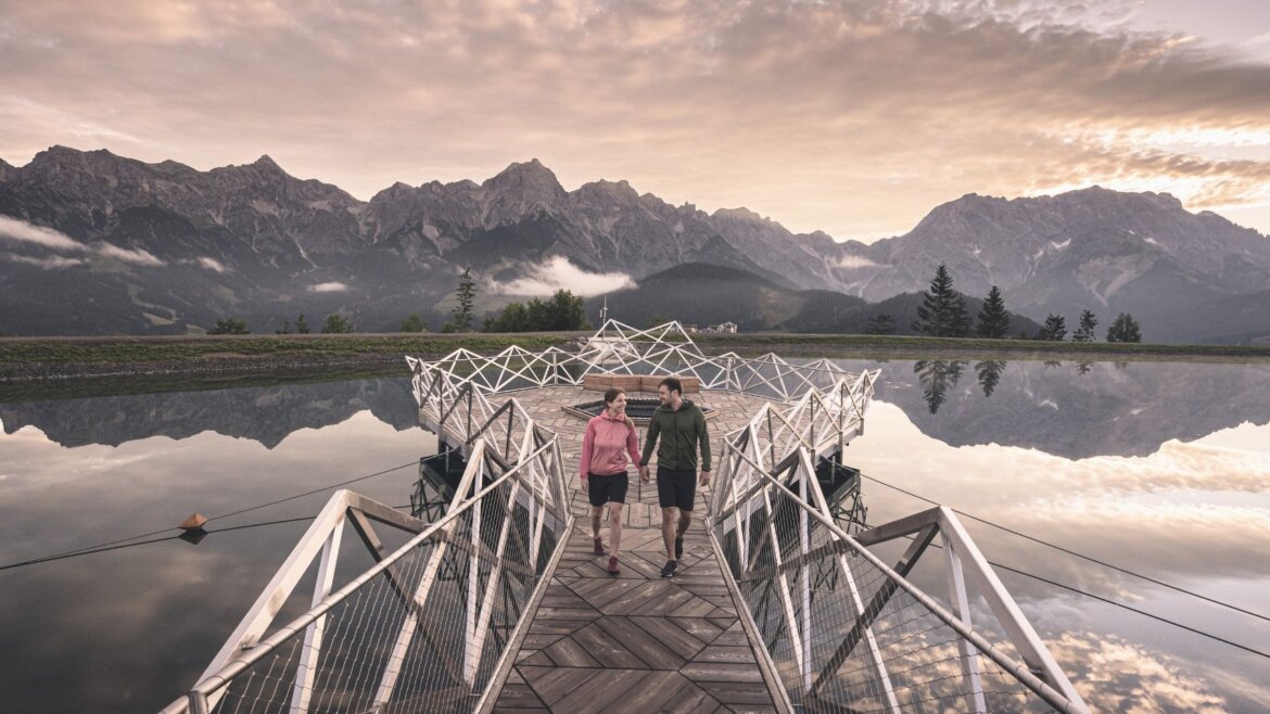 Zwei Menschen gehen über eine geometrische Brücke über stilles Wasser, Berge und die dramatischen Wolken von Die Hochkönigin im Hintergrund. (vergrößerte Ansicht)
