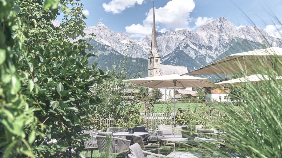 Außencafé mit Sonnenschirmen, Grünflächen und Blick auf die Hochkönigin und einen Kirchturm in der Ferne. (vergrößerte Ansicht)