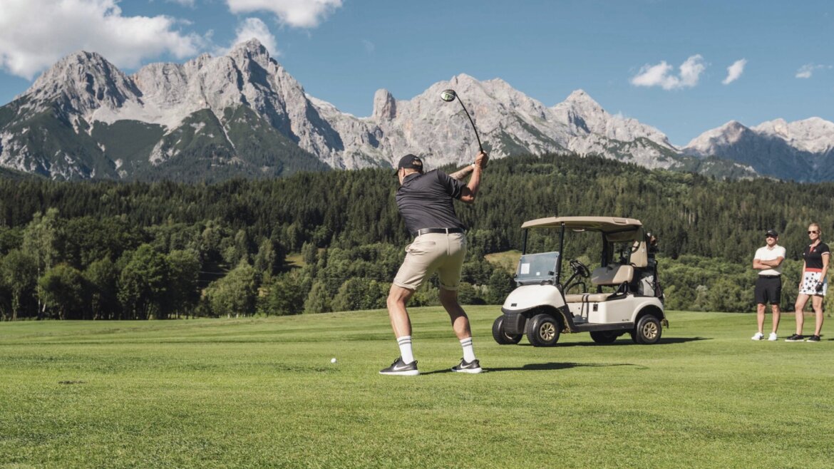 Ein Golfer schwingt auf einem grünen Platz in der Nähe von Die Hochkönigin, mit zwei Beobachtern und Bergen im Hintergrund. (vergrößerte Ansicht)