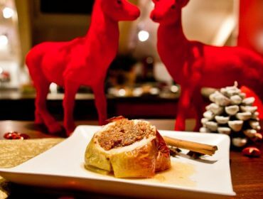 Ein gefüllter Bratapfel auf einem Teller, mit roten Rentierfiguren und einem Tannenzapfen im Hintergrund.