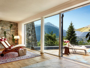 Sonnige Lounge und Terrasse im Schloss Mittersill mit Bergblick, Liegestühlen und offenen Glastüren in einem rustikalen Raum.