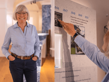 Eine lächelnde Frau steht in einem Raum und zeigt auf eine historische Zeitleiste an der Wand.