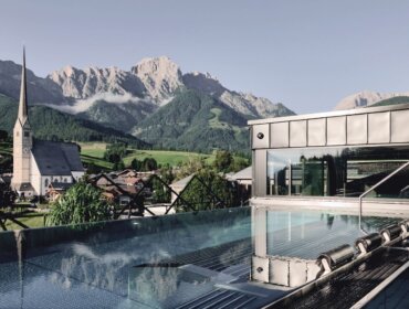 wellnesshotel-hochkoenig-infinitypool-maria-alm.jpg Infinity-Pool mit Blick auf die Hochkönigin, ein malerisches Bergdorf mit einer Kirche und Alpengipfeln dahinter.
