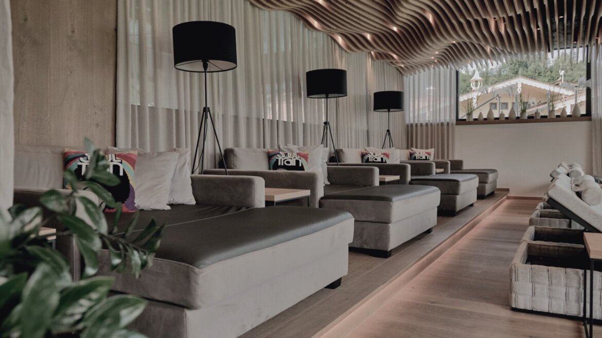 Moderne Lounge in der Hochkönigin mit grauen Chaise-Lounges, Stehlampen, gewellter Holzdecke und üppigen Pflanzen an den Fenstern. (vergrößerte Ansicht)