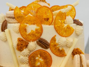 Runder Kuchen, belegt mit getrockneten Orangenscheiben, Macarons, Pekannüssen und Sahne, daneben eine frische Orange.